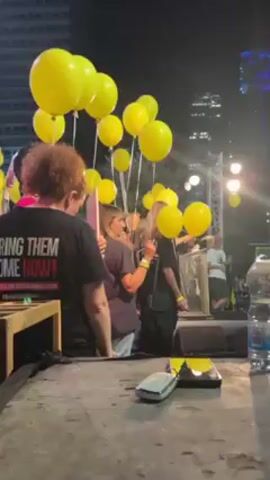 The video shows a crowd of people, mostly women, at an outdoor event at night. They are holding yellow balloons. In the foreground is a stage with some equipment. There is a woman standing at a podium on the stage. Behind her is a large screen displaying an image of a woman. The people in the crowd appear to be listening to the speaker. Some of the people are wearing shirts that say "BRING THEM HOME NOW!" There are tall buildings in the background. The speaker is saying "But our mission is not yet complete. 18 of our..."
