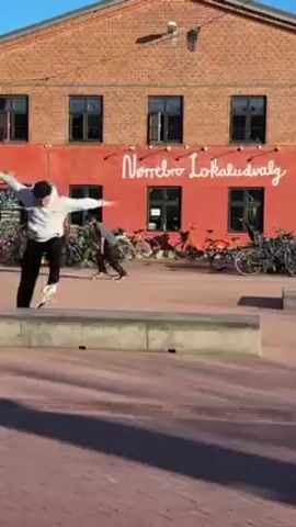 The video shows two people skateboarding in an outdoor urban setting. The first person, wearing a light-colored hoodie and dark pants, attempts a trick on a low concrete ledge but falls off the skateboard. Subsequently, a second person, wearing a dark hooded sweatshirt and dark pants, successfully performs a trick on the same ledge. The location features a brick building in the background with the words "Nørrebro Lokaludvalg" painted in white. Numerous bicycles are parked near the building. The ground is paved with red bricks and the sky is clear.