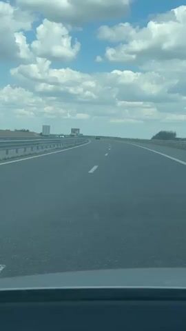 A car is driving down a highway with a blue sky and white clouds above. The road is empty except for a few other cars in the distance. There are billboards on the side of the road. The camera is positioned inside the car, looking forward.