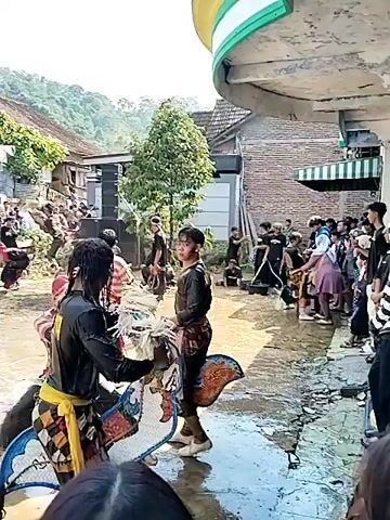 The video shows a performance taking place in what appears to be a village. Multiple individuals are engaged in a traditional dance, wearing costumes. The ground is wet, suggesting it might have rained recently. A crowd is gathered to watch the performance, and several buildings are visible in the background, indicating that the event is taking place in a residential area. There is also sound equipment set up, implying the performance is accompanied by music. The presence of lush greenery and hills in the background suggests that the location is in a relatively rural setting.