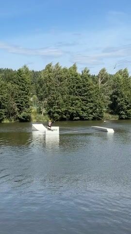 The video shows a wake park on a lake. There are several obstacles in the water, including ramps and a straight rail. A person is wakeboarding and attempting to perform a trick on one of the obstacles. They appear to be going over a ramp and are in the process of falling into the water. The weather is sunny with a few clouds, and the surrounding area is covered in trees.
