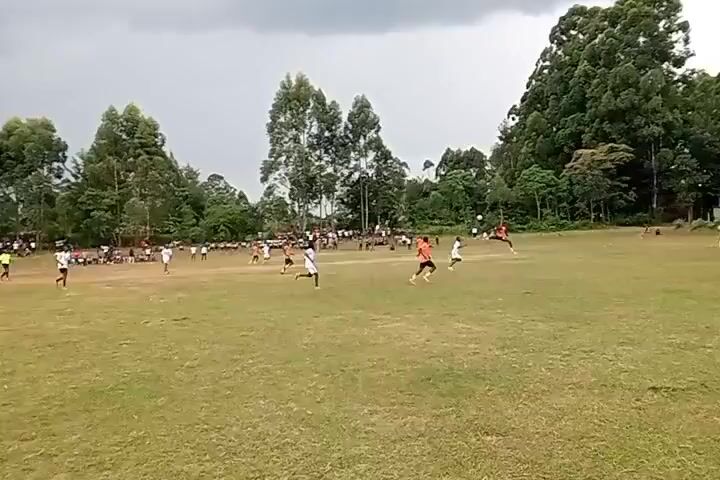 The video shows a soccer game in progress on a grass field. There are many players on the field wearing either white or orange jerseys. Spectators are visible on the sidelines. The weather appears overcast. The field is surrounded by trees. The game is in motion with players running and kicking the ball.
