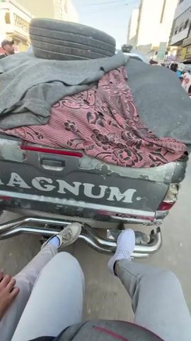 This media file shows a rear view of a pickup truck with a spare tire and blankets loaded in the bed. The truck has "MAGNUM" written on the tailgate in large white letters. Two people's legs are visible in the foreground, suggesting they are either sitting on the tailgate or standing very close behind the truck.

The truck is in a street scene, with buildings lining both sides of the road. In the background, there are other people walking and what appears to be a marketplace with stalls and merchandise. The buildings suggest an urban environment. The sky is clear and blue, indicating a sunny day. The time of day appears to be late morning or early afternoon based on the sunlight and shadows.

Visible text includes "MAGNUM" on the truck and some signage on the buildings in the background, though it is difficult to read clearly. One sign on a building has Arabic script. There is also a partial view of "MITSUBISHI MEGA MAGNUM" above the "MAGNUM" text on the tailgate.

The overall impression is a snapshot of everyday life in a busy urban area, likely a market or commercial street. The presence of the loaded truck and the casual positioning of the people suggest it might be related to transport of goods or people, or simply a moment of rest during a journey through a bustling town.