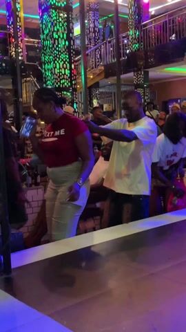 The video shows a woman in a red shirt that says "TEXT ME" and light-colored pants dancing at what appears to be a nightclub or bar. A man in a white shirt is standing behind her and holding her, also dancing. Other people can be seen in the background. The setting appears to be indoors with colorful lighting and architectural features.
