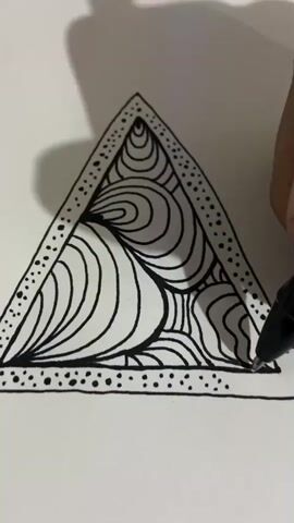 A close-up video shows a person's hand holding a black marker and drawing on a white piece of paper. The paper contains a large triangle outline filled with intricate black and white line art. The lines are organic and swirling, creating a mesmerizing pattern within the triangle. The drawing is nearly complete, with the marker actively adding the final details to the bottom right corner of the triangle's interior. The background is a plain white surface, with a subtle shadow cast from above, suggesting overhead lighting.