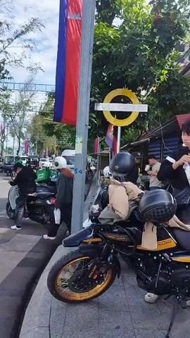 The video pans across a street scene with several motorcycles parked along the sidewalk. In the foreground, a black and yellow motorcycle is prominently displayed, equipped with helmets and bags. Two young men are interacting near this motorcycle; one is holding a drink and appears to be handing something to the other, who is wearing a light blue shirt and holding a black helmet. Further down the street, other motorcycles, including a white scooter and a red and black scooter, are visible. People are walking and standing on the sidewalk, and a business with an outdoor seating area and umbrellas is present. The environment appears to be urban, with buildings and power lines visible. A Cambodian flag is also visible on a pole. The overall impression is a lively street scene with a focus on motorcycles.