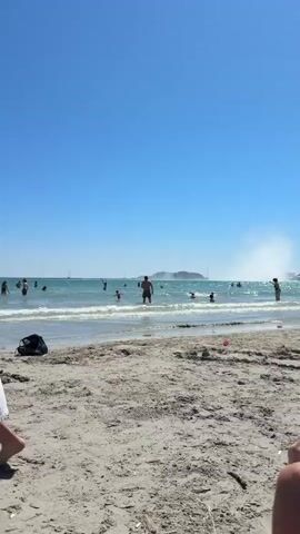 The video captures a bright, sunny day at a bustling beach with clear blue skies. The sea is calm, with turquoise water visible in the background.

The primary subject is a young boy, wearing a light-colored cap, a white robe, and patterned swim shorts, who runs barefoot from left to right across the sandy beach.

In the initial frames, two children are seen playing in the sand near the water's edge, utilizing sand buckets and shovels. Several other people are visible swimming or wading in the ocean further out.

As the camera pans, more of the beach comes into view. Numerous individuals are relaxing on lounge chairs and beach towels, some under umbrellas. Palm trees are scattered along the beachfront, and buildings are visible in the distance, suggesting a resort or town area. A soccer ball rests on the sand near some lounge chairs.

The main activity shown is the boy running, while other people engage in typical beach activities such as sunbathing, swimming, and playing. The scene depicts a lively and sunny beach environment. Briefly, a sign on a building in the background appears to display the text "LA VAGUE".