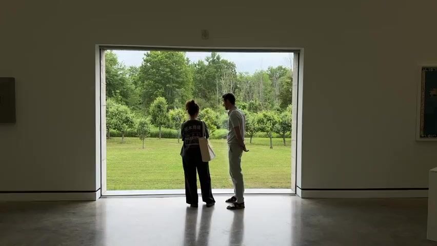 The video shows two people standing in front of a large window in a gallery. The window looks out onto a grassy field with trees. The person on the left is a woman with her back to the camera, wearing a dark shirt and pants and carrying a tote bag. The person on the right is a man wearing a light-colored shirt and pants. They are looking out the window. The woman turns and walks towards the camera, and the man follows. The video appears to be a short, static shot with no sound.