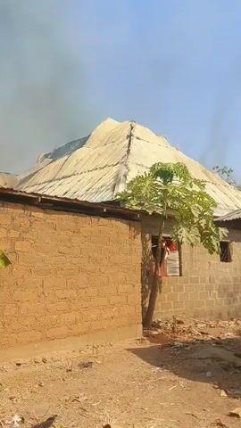 The video shows smoke rising from the roof of a building made of brick and corrugated metal. The building is surrounded by a dirt yard and a brick wall. There is a small tree in front of the building. The sky is blue and there are no people visible in the video.