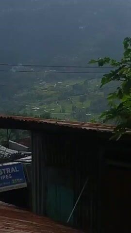 A short, vertical, medium-shot video shows a view of a valley from a hillside. In the foreground, the corrugated metal roof of a building is visible. To the left, a blue and white sign for "ASTRAL PIPES" is attached to the building. The valley below is a patchwork of green fields and scattered houses, with a road winding through it. The background is dominated by misty, tree-covered hills. Sunlight filters through the foliage, creating dappled shadows. The scene is tranquil and serene, with a sense of rural remoteness.