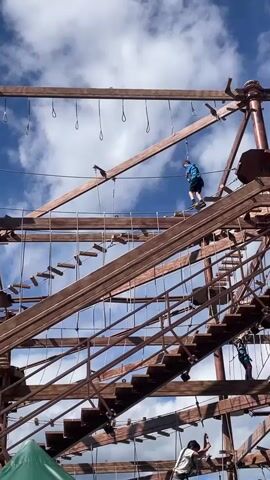 The video shows a high ropes course with multiple levels of wooden platforms and rope bridges under a blue sky with white clouds. Several people are navigating the course. In the upper part of the frame, a young person in a blue shirt and dark shorts is walking on a narrow wooden plank bridge, attached to safety lines. Below them, a series of suspended wooden steps are visible. Further down, another person in a dark shirt and shorts is seen descending a wooden staircase. Near the bottom of the frame, a person wearing a white top is holding onto a rope, presumably on a lower level of the course. The ropes course is constructed of brown wooden beams and metal supports. The overall environment appears to be an outdoor adventure park.