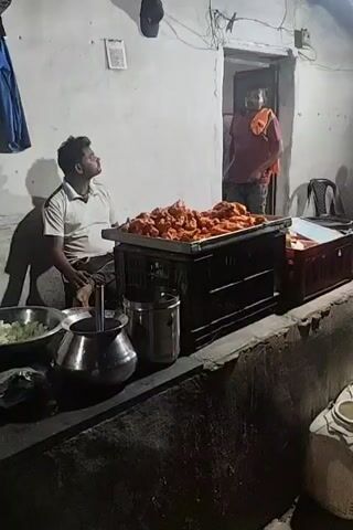 The video shows two men in what appears to be a kitchen or food preparation area at night.  The first man, wearing a light-colored polo shirt and dark shorts, is seated on the left side of the frame, looking upwards and to his right.  In front of him is a large metal tray piled high with pieces of what looks like fried chicken or meat. To his left are several large metal bowls, one containing a green leafy vegetable.  To the right of the meat tray is a dark-colored crate.  In the background, a doorway is visible, and a second man, wearing a red shirt and dark pants, is standing in the doorway, looking towards the viewer.  He has his arms crossed.  The walls of the room are a light color and appear to be made of concrete or plaster.  There are shadows cast on the wall behind the seated man.  The overall lighting is dim, suggesting it is nighttime.  The second segment shows a young man wearing a black sweatshirt with white lettering and dark pants. He is standing in front of several large cooking pots, one of which has a metal lid. On top of one of the lids is a plate with a small amount of food and a metal utensil. He appears to be looking to his right.  The third segment shows a similar scene with the young man in the black sweatshirt standing with his hand on his hip.  He is looking to his right.  Two large pots are visible in front of him, with lids. On one lid, there is a plate and a small metal container.  The fourth segment shows a close-up of a large pot with a lid, and a metal utensil resting on top of the lid.  The background is blurred.