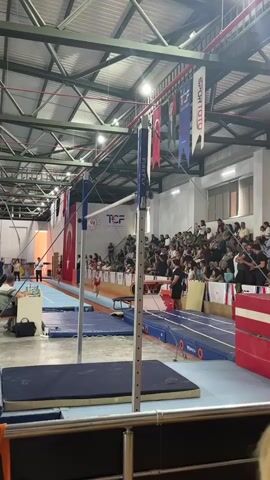 The video shows a gymnastics competition. In the foreground, there are blue mats and a gymnastics apparatus. In the background, there is a crowd of spectators sitting in bleachers, watching the competition. A female gymnast, wearing a red leotard, is performing on a blue mat with white and red markings, which appears to be a vaulting runway. She is in the process of a run and approach to a vaulting table, which is out of frame. There are flags hanging from the ceiling of the gymnasium, including Turkish flags and banners with text like "SPOR TOTO" and "TCF". The environment is an indoor gymnasium with a metal roof structure and lighting fixtures.