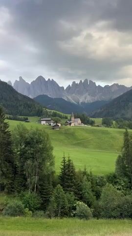 A drone shot pans across a pastoral scene in a valley, with the jagged peaks of the Dolomites mountains in the background. The foreground is dominated by lush green fields, dotted with scattered trees and shrubs. In the midground, a small cluster of buildings, including a church with a steeple, is nestled on a gentle slope. The sky is overcast with dramatic, gray clouds. The overall impression is one of serene, natural beauty.