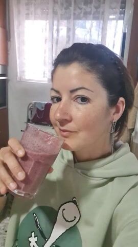 A woman takes a sip from a glass of dark pink smoothie, holding the glass in her right hand. She looks at the camera with a slight smile. She's wearing a light green hoodie with a cartoon print on the front, and silver chain earrings. She has dark hair pulled back with a braided headband, a nose ring, and some freckles. The background includes a window with a sheer, patterned curtain, and a glimpse of furniture and other items in a room. The lighting is bright.