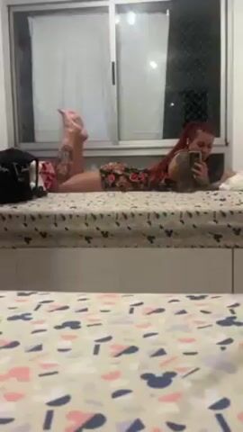 A woman with red hair and tattoos on her legs and arm is lying on a bed. She is wearing a floral dress. She is holding a cell phone in her hand and looking at the screen. The bed has a Mickey Mouse-themed bedsheet. A window is behind the bed. A backpack and a pair of socks are on the bed next to her. The room is a light color.