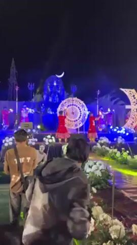 People are watching a dance performance at night. The dancers, wearing red and yellow costumes, are on a stage decorated with white flowers and illuminated by blue lights. Behind them is a large statue of a face, possibly a deity, also lit in blue. A crescent moon decoration is above the statue. There is a large circular decorative object on the stage with the dancers. Several people are filming the performance with their phones.