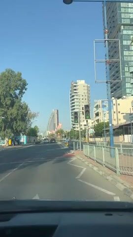 The video shows a view from inside a vehicle driving on a street. It is a sunny day with a clear blue sky. On the right side of the road, there is a tall modern building with many windows. Further down the road, there are other buildings, some of which are also tall and modern, with curved architectural designs. There are traffic lights, street signs, and a pedestrian crossing visible. Trees line the left side of the road. The street is paved and has lane markings. Cars are driving in the same direction as the viewer. The overall impression is of a city street in a developed area.