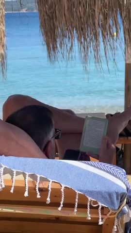 A person is lying on a beach lounger under a thatched roof umbrella. They are wearing sunglasses and reading an e-reader. A blue and white striped towel with tassels is draped over the lounger. The ocean is visible in the background, with clear blue water and a sandy beach.