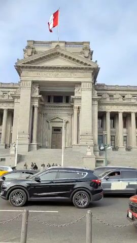The video shows an outdoor scene with a line of cars and a light-colored wooden coffin placed on a stand. The coffin is decorated with floral arrangements in pastel colors. In the background, there is a large, stately building with columns and a pediment, labeled "Palacio de Justicia." A person can be seen driving one of the cars. The video is shot from a ground-level perspective and pans slightly to follow the line of cars. The scene appears to be taking place during the daytime under a partly cloudy sky. There are pedestrian barriers in front of the coffin and the road. Someone is talking, but the conversation is hard to understand.