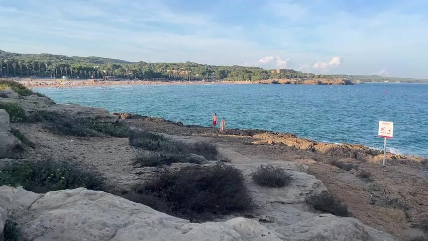 The video shows a coastline with rocky shorelines and a calm blue sea under a partly cloudy sky. In the distance, a crowded beach is visible with many people enjoying the sun and water. Closer to the foreground, two individuals are standing on the rocky shore. A signpost is planted in the rocks. Further along the coast, another individual is seen fishing with a fishing rod. The overall scene depicts a serene coastal landscape with recreational activities.