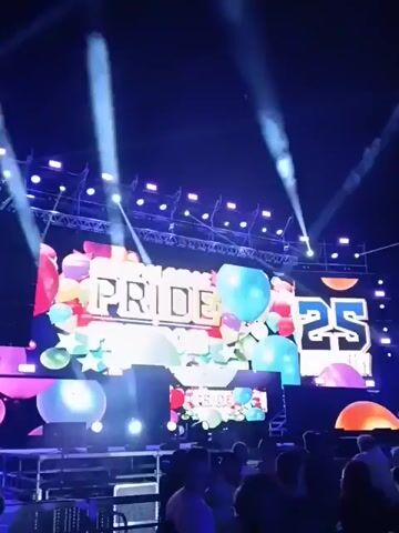 The video shows a nighttime outdoor event, likely a concert or festival, in a crowded space with a large stage. The stage features a brightly lit screen displaying the words "PRIDE CELEBRATION" with colorful balloons and the number "25". Above the stage, several spotlights shine upward into the night sky, illuminating it with beams of white and blue light, and smoke effects. A large crowd is visible in front of the stage, mostly standing and facing the stage. Buildings with lighted windows are visible in the background behind the crowd, indicating an urban setting. The overall atmosphere is lively and festive.