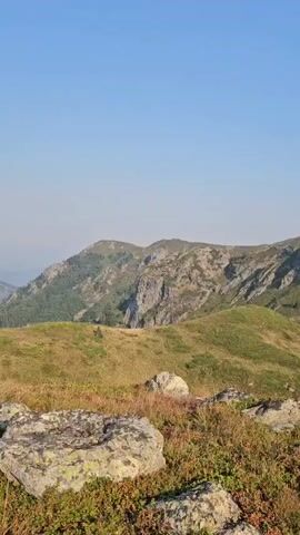 The video pans across a vast mountain landscape on a clear day. The foreground features rocks and low-lying green and red foliage. The middle ground reveals rolling green hills dotted with patches of sparse trees and rocky outcrops. In the background, majestic mountains rise, some with exposed rock faces and others covered in vegetation. The sky is a clear, bright blue. The camera slowly moves from right to left, revealing more of the expansive mountain range and the valleys between peaks.