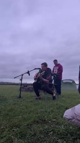 A man sits on a folding chair outdoors, playing an acoustic guitar and singing into a microphone. He is wearing a black jacket with red accents and dark pants. Another man stands behind him, facing away from the camera, wearing a purple hooded sweatshirt. A green tent is visible in the background, along with a body of water. The sky is overcast, and the ground is covered in grass. The video appears to be taken at dusk or during a cloudy day. There are also some orange ropes or markers on the ground in the foreground.