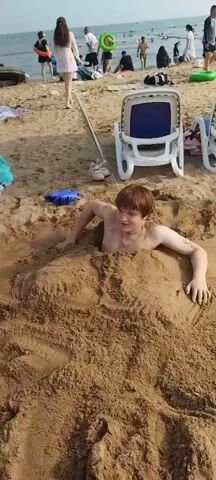 This is a video taken on a sandy beach, likely during the daytime under clear weather conditions. The main subject is a young male who is partially buried in the sand. He is shirtless and appears to be struggling to free himself from the sand, with his arms and legs submerged. He has a playful and somewhat determined expression on his face, suggesting this is a fun activity rather than a genuine emergency.

In the background, the beach is populated with other people, some of whom are walking or sitting. There are beach chairs, umbrellas, and a glimpse of the ocean with calm water. The presence of lounge chairs and numerous beachgoers indicates a popular tourist or recreational area. The overall atmosphere seems relaxed and summery, consistent with a beach day.

Specific objects visible include a pair of pink children's shoes lying on the sand, a blue inflatable object, and two white plastic beach chairs with blue seats. The sand itself is a prominent element, forming a mound around the boy. The horizon shows the sea meeting the sky under what appears to be a bright sky, implying it's likely late morning or afternoon. There is no visible text in the video. The location context of Qinhuangdao, China, suggests this is a beach in that coastal city.