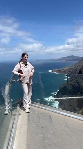 The video shows several people at a viewpoint. At the beginning of the video, a young woman stands leaning against a glass barrier looking at the ocean. She is wearing a tan pantsuit. The next person, a young man, looks over the edge. An older woman and man look at a cell phone. The man is wearing a white t-shirt, a backpack, and a baseball cap. The viewpoint overlooks a body of water and mountainous terrain. The sky is blue with clouds.