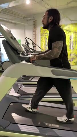 A man with tattoos on his arms and face walks on a treadmill. He is wearing glasses, a black t-shirt, and black pants. The treadmill is in a gym with other treadmills visible in the background. There is a picture of trees on the wall. The man raises his hand and makes a gesture with his fingers.