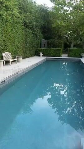 The video shows a lush, green backyard with a swimming pool. The pool is surrounded by white tile and hedges.  Beyond the hedges, there is a patio area with people sitting at tables. There are several people seated around a table, some standing nearby, and others entering the house through an open door. The house is white with multiple windows and brick chimneys. The weather appears sunny.