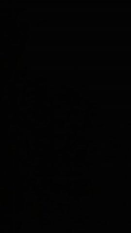 A person wearing a traditional Arab headscarf and garment stands in front of a dark background, singing with their eyes closed. The person's mouth is open as they sing. The lighting is dim.