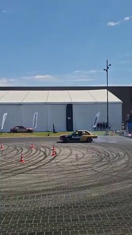 The video shows a car drifting in a parking lot or asphalt area. There are orange traffic cones set up in a circular pattern. The car is black with red accents and appears to be a sedan. Smoke is coming from the rear tires of the car as it drifts. In the background, there is a large white tent structure and a blue sky with some clouds. There are other cars and people visible in the distance.