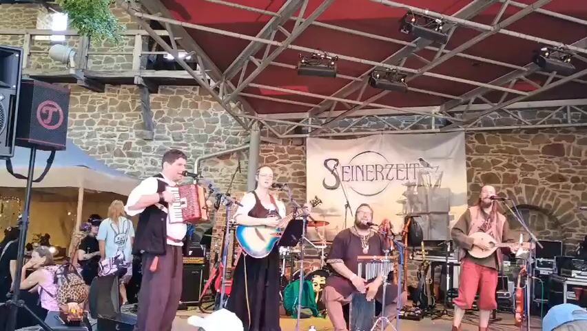 The video shows a live performance by a band called "Seinerzeit" on an outdoor stage.  There are four musicians visible on stage. On the far left, a man is playing an accordion and singing into a microphone. Next to him, a woman is playing an acoustic guitar and singing. In the center, a man is playing a xylophone-like instrument and singing. On the far right, a man is playing a stringed instrument, possibly a lute or mandolin, and singing. The band is performing under a red canopy supported by a metal structure. Behind them, a banner with the band's name and a stylized ship is displayed. There are speakers and sound equipment on the stage. In the background, a stone building and tents are visible, suggesting a festival or outdoor event. Several people are in the audience, some standing and others sitting. The atmosphere appears to be lively and festive.