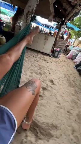 The video shows a person lying in a hammock on a sandy beach. The subject's legs are visible, with a butterfly tattoo on their left thigh and an anklet on their right ankle. The hammock is green and appears to be suspended from a wooden structure, possibly part of a beachside bar or cafe. In the background, there are several green coconuts in a basket, a white cabinet with items on top, and other beachgoers in the distance. The scene suggests a relaxed, tropical atmosphere, likely during daylight hours given the bright lighting. The weather appears to be clear and warm, suitable for a beach outing. The overall mood conveyed is one of leisure and enjoyment of a vacation or a day at the beach.