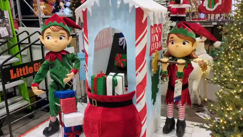 The video shows a Christmas display in a store. Two elf figures, one male and one female, stand on either side of a booth labeled "Santa's Toy Factory." The male elf is dressed in green and holds a string of lights, while the female elf is in red and holds a hammer. A red bag with gifts is in front of the booth. The booth has a blue and white roof resembling snow. A small Christmas tree is on the right side. A ladder with the text "Little Giant" is visible in the background.