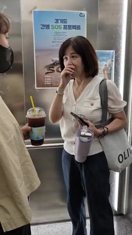 Two women are in an elevator. The woman on the left is wearing a tan top and a black mask. She is holding a cup of iced coffee with a yellow straw. The woman on the right is wearing a white v-neck shirt and blue jeans. She has dark short hair and is wearing a black mask. She is holding a smartphone in her left hand and a purple tumbler in her right hand. She is looking towards the man on the left and has her right hand near her mouth. A poster is visible on the elevator wall behind them.