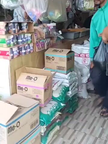 The video pans across the interior of a small shop. The shelves are stocked with various products, including what appear to be canned goods and bags of rice or pet food. In the foreground, a man wearing a teal long-sleeved shirt and dark pants walks past, carrying a black plastic bag. He has a dark complexion and a mustache. Further into the store, another man in a brightly colored shirt and white shorts is visible, also with a black bag. In the background, there are stacks of bags, some green and red, with labels like "Mister Puss" and "Cat Hoize." There are also green propane tanks and a white birdcage. The overall impression is of a general store or a pet supply shop.