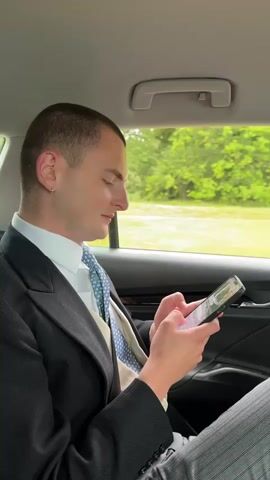 A man in formal wear, consisting of a dark suit jacket, a white collared shirt, a blue patterned tie, and a light-colored waistcoat, is seated in the back of a car. He has short, dark hair, an earring in his left ear, and is looking down at a smartphone held in both hands, his thumbs positioned as if typing or scrolling. He then looks up and smiles at the camera, before the camera pans down and focuses on the blurred green landscape passing outside the car window, indicating movement.