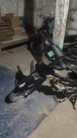 The video shows a dimly lit indoor scene with several motorcycles. In the foreground, a black motorcycle is leaning against a pole. Its front wheel is turned to the left, and the engine area is exposed, with wires and mechanical parts visible. The seat is black with a wavy pattern. The rear wheel is visible, and a red shock absorber is present. The motorcycle is partially covered by a blue and white tarp. In the background, another black motorcycle is parked, with its license plate clearly visible as S 3032 BD. There are cardboard boxes stacked on a wooden shelf to the left of the frame, and a bucket with some items inside is visible in the far background. The walls appear to be made of wood and are painted with some peeling blue and white paint. The floor is concrete.