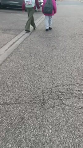 The video shows two children walking away from the camera on an asphalt road. They are both wearing backpacks and appear to be heading away from a car parked on the side of the road. The environment appears to be a residential street with houses and trees visible in the background.