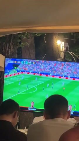 The video shows a screen displaying a soccer match. The score is Belgium 0, Senegal 1. The time is 45:13. There are several people watching the screen from behind. The screen is large and is mounted on a wall or stand. The environment appears to be an indoor space with some outdoor foliage visible in the background. The lighting is dim, suggesting it might be nighttime or an indoor venue. The focus is on the soccer match being broadcast.