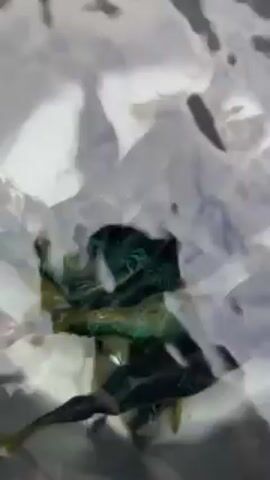 The video shows a close-up, somewhat obscured view of what appears to be a dark-colored object surrounded by foamy, agitated water. The object is partially submerged, and its exact nature is unclear due to the poor image quality and movement of the water. There are suggestions of green and brown colors within the object. The water is mostly white and gray, giving the impression of a turbulent or rapidly flowing environment.