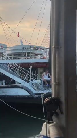 A man sits on a small stool on the deck of a boat. He is wearing a white tank top and white shorts. The boat is docked next to a larger boat with multiple decks. The sky is cloudy and appears to be dusk. There are other boats and structures visible in the background.