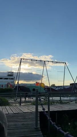 The video shows a public playground at sunset.  A person is seen from the back, walking towards the playground, which features large swings and a climbing structure. Several children are on the swings and climbing structure. The sky is blue with some clouds, and the sun is setting in the background. The playground is built over a shallow pond, and wooden walkways connect the various features.