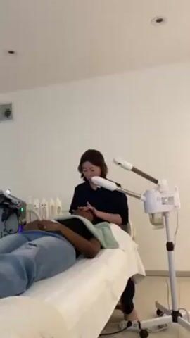 In a brightly lit indoor setting, a light-skinned esthetician with short dark hair is performing a facial massage on a darker-skinned person lying on a treatment bed. The esthetician is wearing a dark-colored top and is seated to the side of the bed, gently massaging the forehead and face of the person receiving the treatment. The person on the bed is wearing a black top and blue jeans and has a green towel draped around their neck. A white facial steamer is positioned near the head of the bed, and several bottles of skincare products are visible on a table in the background, along with a machine with a blue light. The treatment room appears clean and minimalist, with white walls and a tiled floor.