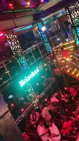 The video provides an elevated, panning view of a bustling, multi-level indoor entertainment venue, likely a bar or lounge, at night. The scene is illuminated by an array of artificial lights, including vibrant multi-colored LED strips on decorative pillars and along the ceiling, and warm string lights suspended above the main seating areas.

A prominent green LED sign displaying "Heineken" is visible on the wall of the ground floor, serving as a focal point. The venue features a ground floor with numerous tables and seating arrangements, and an upper balcony level, both filled with patrons. A stylish staircase with warm, golden lighting connects the two levels.

Many people are present, seated at tables and in lounge-style seating, engaging in conversation and socializing. Drinks in cups and bottles are visible on several tables. The individuals appear to be adults, dressed in casual attire, and seem relaxed and enjoying the lively atmosphere. The video slowly pans across the ground floor, offering different perspectives of the crowded space and the overall ambiance. The setting suggests a popular nightspot or social gathering place.