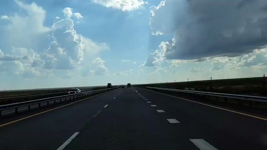 The video is a first-person perspective, likely from a vehicle, traveling on a multi-lane highway under a bright, cloudy sky. The road is asphalt with clear lane markings, including solid yellow lines on the right and dashed white lines in the center. Guardrails are visible on both sides of the road, with a flat, grassy landscape stretching out to the horizon. Several vehicles are visible in the distance, including trucks and cars, all traveling in the same direction. The sky is dominated by large, white cumulus clouds with some darker storm clouds forming on the right side, and shafts of sunlight breaking through. The overall impression is one of a long, open road under a dynamic sky, suggesting a journey through a vast, possibly rural, landscape.