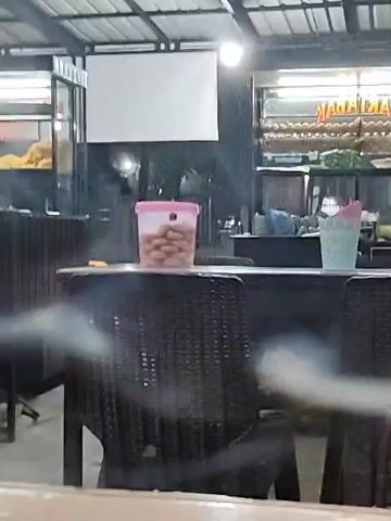 The video presents an outdoor dining area under a metal roof structure, likely a restaurant or food stall. The focus is on a table and chairs, with a plastic container filled with small, round food items on the table's surface. Behind the table, there are food display areas with various items, suggesting a place serving a variety of dishes. There's a blank white screen or sign on the back wall. The scene appears to be at night, lit by overhead lights. The sound of a vehicle passing by is clearly audible.